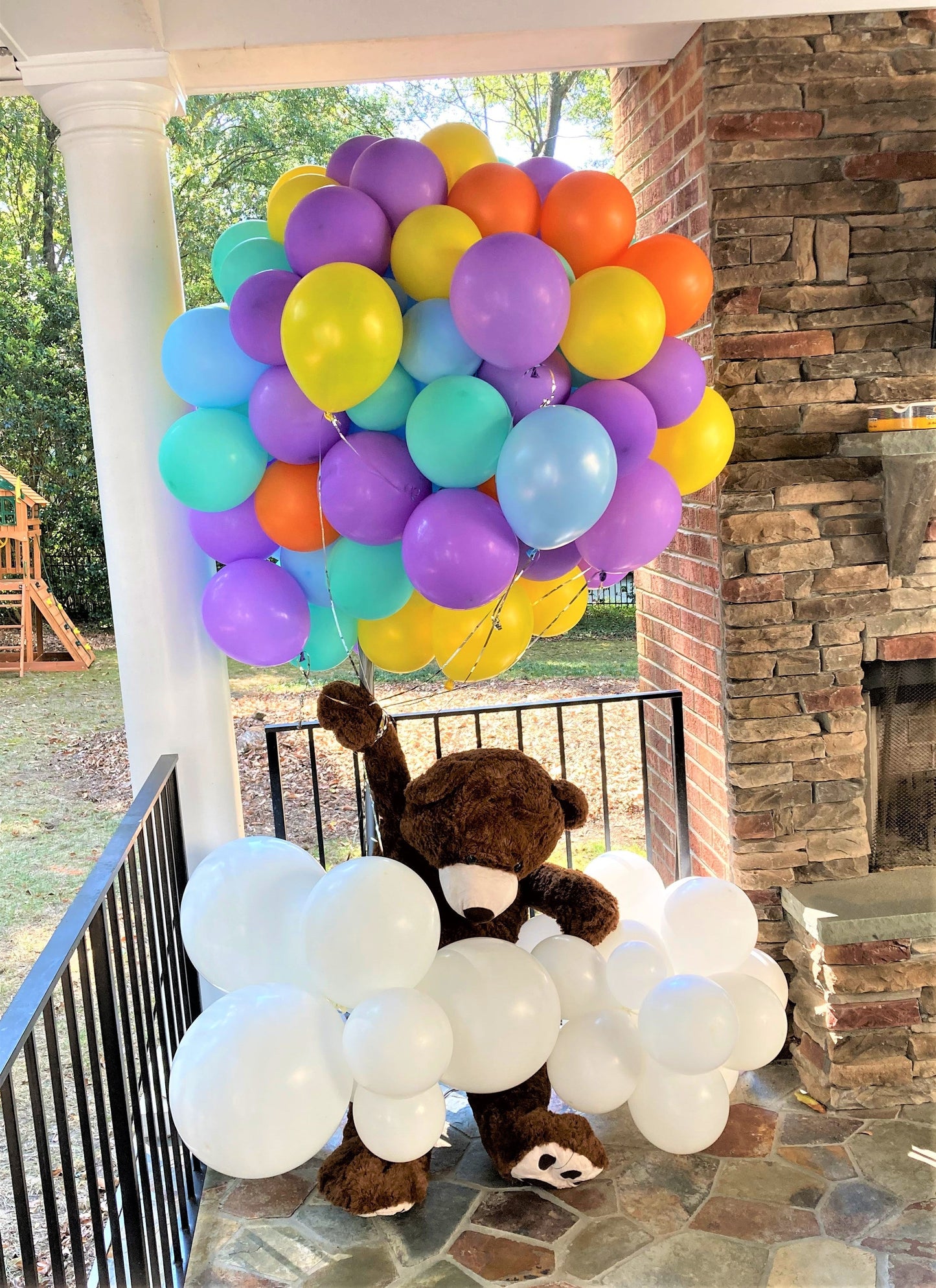 bunch of balloons yellow, lavender, light blue and orange with floating teddy bear attached wrapped in a white cloud