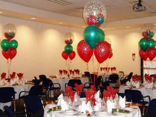 A Christmas-themed balloon centerpiece featuring round and oversized balloons in red, green, and clear colors with bubble decorations, arranged in a festive setting with tables and decorations.