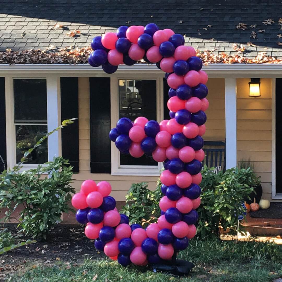 large balloon sculpture  number 3 made with pink and purple balloons on a lawn in front of a house.