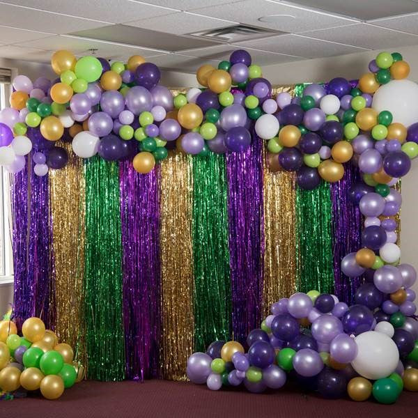 A Mardi Gras-themed balloon garland featuring a variety of green, gold and purple balloons and a gold, green and purple-fringed backdrop.