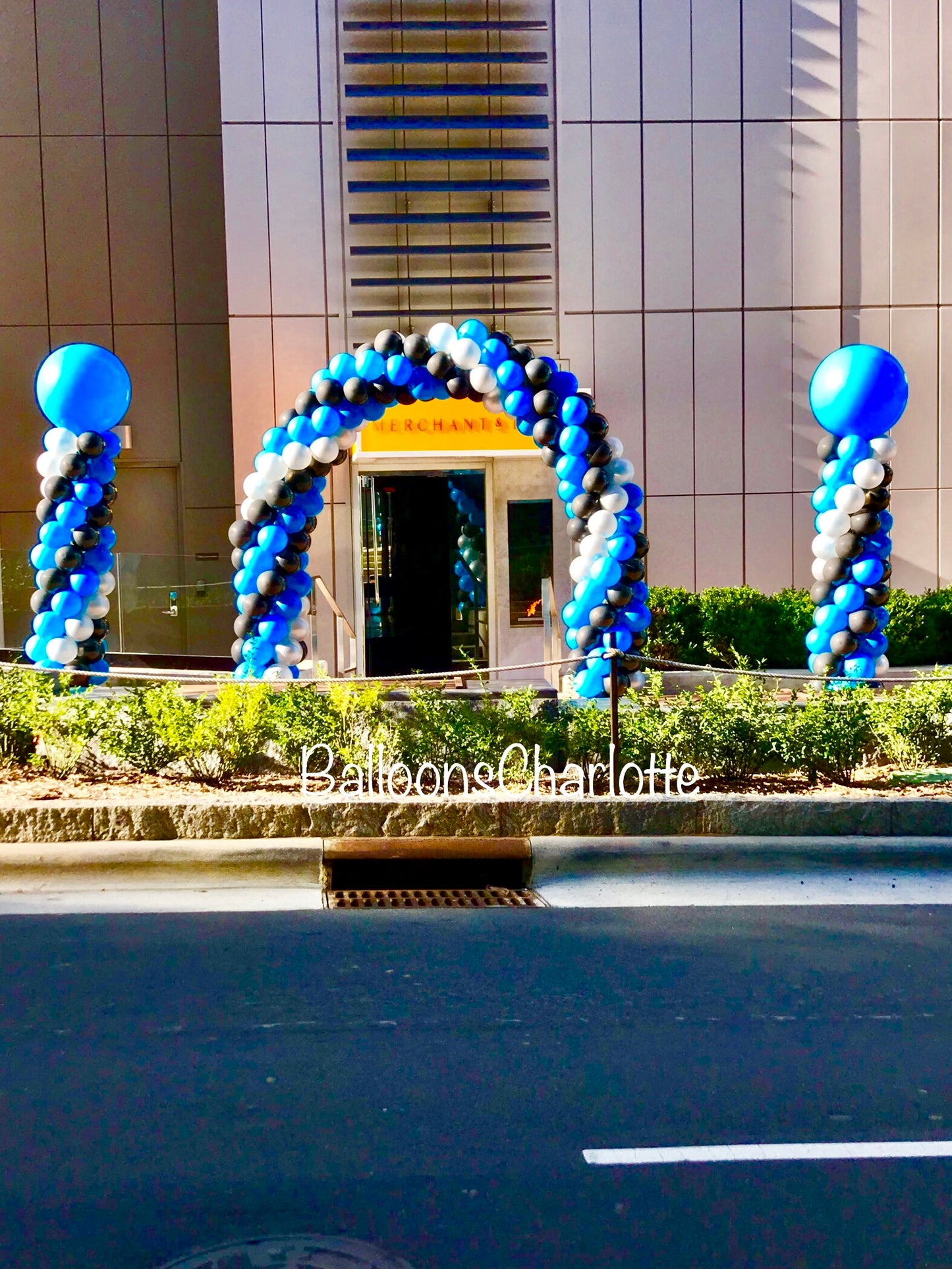 A blue, silver and black balloon arch and columns with big blue balloons on top sitting in front of big building.