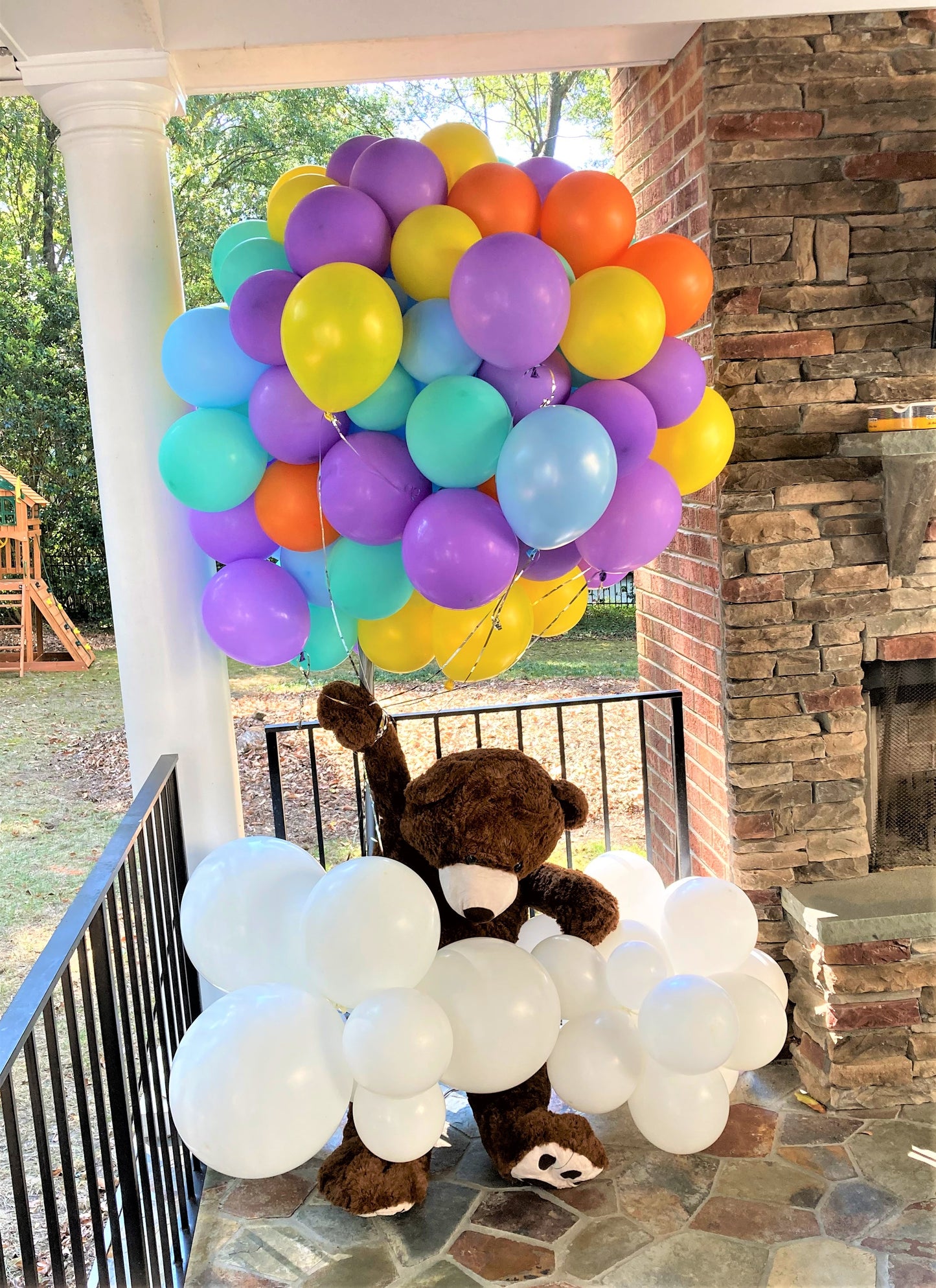 bunch of balloons yellow, lavender, light blue and orange with floating teddy bear attached wrapped in a white cloud