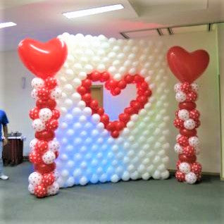 A white balloon wall with an open red heart in the center.  a photo wall with Valentine columns, decorated with red and white balloons with red hearts on top.